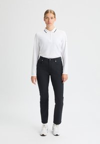 Vit långärmad polo skjorta med svart krage, kombinerad med svarta tajta byxor. Har fickor och silverdetaljer. Modellen bär vita sneakers.