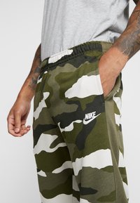 Närbild på en person som bär gröna och vita camouflage Nike-mjukisbyxor, med en hand i fickan, synliga tatueringar på underarmarna och en ljusgrå tröja.