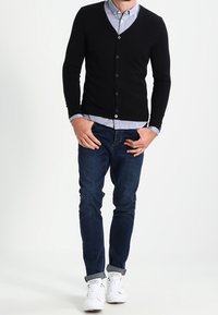 Svart knappad cardigan över en ljusblå skjorta med krage, tillsammans med slim-fit mörkblåjeans och vita sneakers.
