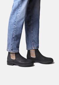 Botas Chelsea de cuero negro con paneles laterales elásticos a rayas, que presentan una suela texturizada y punta redondeada, combinadas con jeans de mezclilla azules.