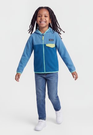 Niño sonriente caminando hacia adelante con una sudadera azul con solapa amarilla en el bolsillo, vaqueros azules y zapatillas blancas sobre un fondo liso.