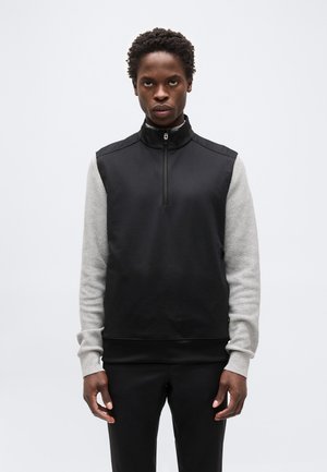 Junger Mann steht und blickt nach vorne, trägt eine schwarze ärmellose Weste mit Reißverschluss über einem grauen, langärmligen, strukturierten Shirt und schwarzen Hosen.