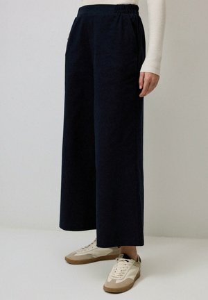 Pantaloni larghi blu navy con una cintura elastica e tasche laterali, abbinati a sneaker bianche con dettagli beige. Tessuto morbido.