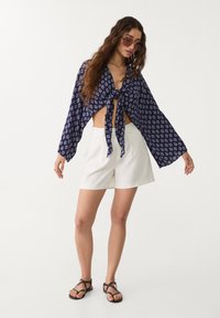 Crop top à motif navy avec des manches larges et un nœud à l'avant, associé à un short taille haute blanc et des sandales à lanières marron.
