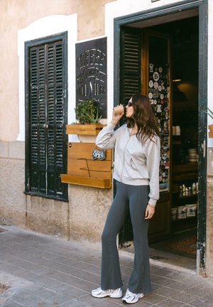 Felpa grigia con zip, leggings grigi a vita alta svasati, e scarpe da ginnastica bianche. In piedi davanti a un caffè con una bevanda, luce solare su superfici texture.