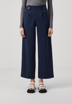 Femme portant un pantalon large bleu marine avec des boutons à la taille, un haut gris en maille côtelée à manches longues, et des sandales blanches.