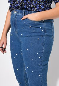 Jeans in denim con vestibilità slim, caratterizzati da numerose perle finte bianche sparse su un tessuto blu scuro. Inclusi tasche laterali e chiusura frontale con bottone.