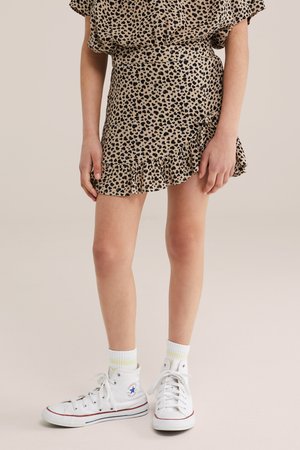 Mladić u bež mini suknji s volanima i leopard uzorkom, odgovarajućoj majici, bijelim čarapama do gležnja i bijelim visokim tenisicama marke Converse.
