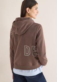 Femme portant un sweat à capuche marron avec « DES ease » brodé dans le dos, superposé à une chemise à rayures bleues et blanches et un jean foncé.