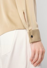 Blusa di seta beige con polsini, caratterizzata da un bottone nero accento e uno spacco laterale, abbinata a pantaloni bianchi. Texture liscia, design elegante.