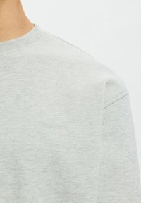 Gros plan sur une personne portant un sweat-shirt gris clair à col rond avec le texte "Selected" subtilement brodé sur la poitrine.