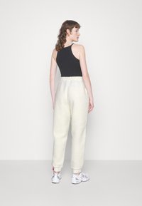 Weiße, flauschige Sweatpants mit einem Kordelzugbund, kombiniert mit einem schwarzen ärmellosen Oberteil. Das Modell trägt helle Sportschuhe. Rückansicht angezeigt.