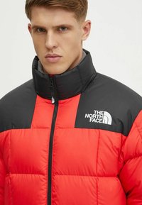Giacca pom pom rossa con colletto e parte superiore nera, dotata di chiusura frontale con zip e logo The North Face bianco sul lato sinistro.