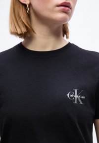 Γυναίκα με κοντά ξανθά μαλλιά που φορά χρυσούς κρίκους στα αυτιά και ένα μαύρο t-shirt Calvin Klein με το λογότυπο στο στήθος.