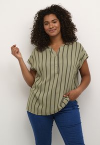 Blouse rayée verte avec un décolleté en V, des manches courtes et un détail de boutons. Tissu lisse avec des rayures verticales dans des tons de vert plus foncé. Jeans visibles.