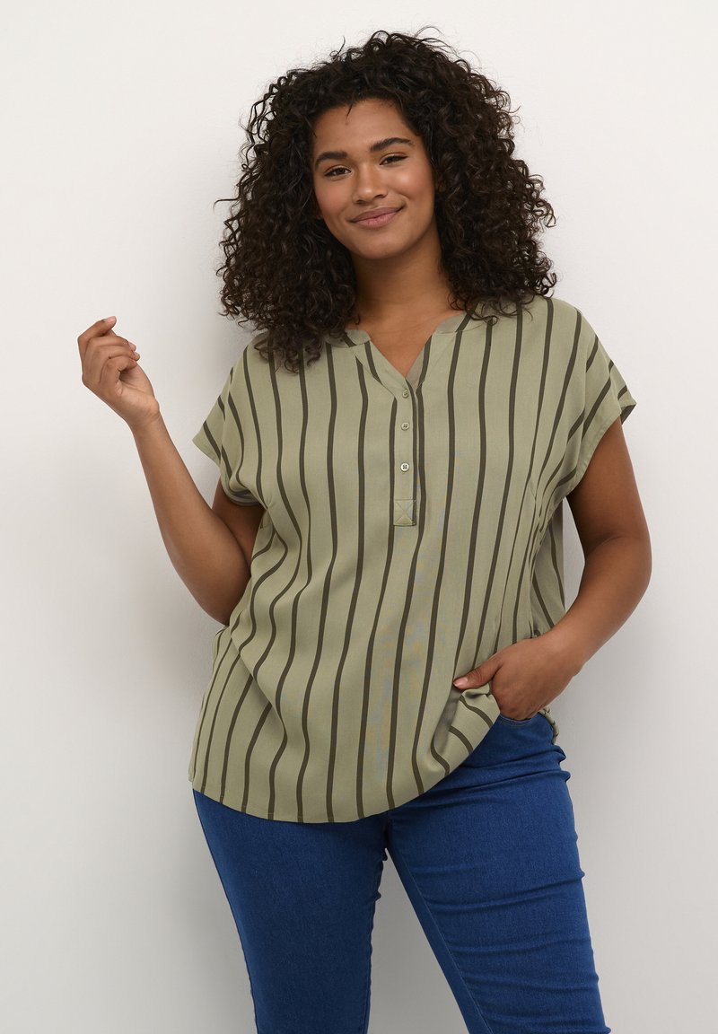 Blouse rayée verte avec un décolleté en V, des manches courtes et un détail de boutons. Tissu lisse avec des rayures verticales dans des tons de vert plus foncé. Jeans visibles.