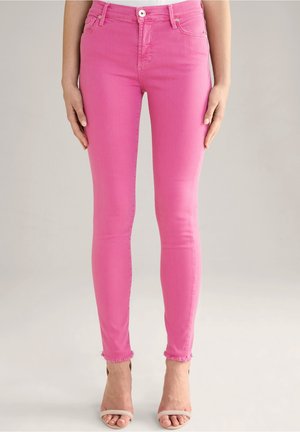 Femme portant un jean skinny rose vif à ourlets effilochés et des sandales blanches à talons hauts et bouts ouverts, debout devant un fond uni.