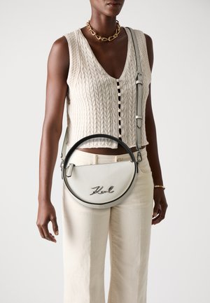 White leather circular handbag with a black trim, featuring a silver logo, worn crossbody with a long strap. Textured knit top, light-colored pants.