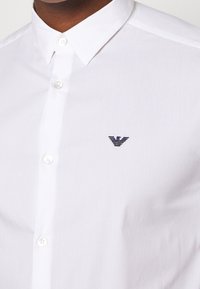 Vit skjorta med knappar och en smal krage. Har en liten marinblå logotyp på vänster bröst och ett slätt, texturerat tyg.