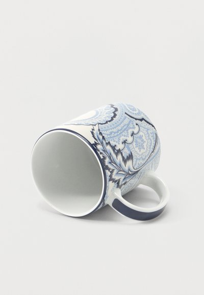 Tasse en céramique blanche avec des motifs floraux et paisley bleus complexes, posée sur le côté avec une anse courbée sur un fond uni.