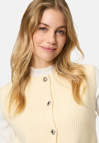 Helles gelbes, geripptes Top mit großen metallischen Knöpfen, getragen über einem weißen Oberteil mit fransigem Ausschnitt. Weicher Stoff und taillierte Passform.