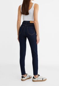Dark blue skinny jeans with a high waist, featuring back pocket stitching and a tan leather label at the waistband. Paired with gray sneakers.