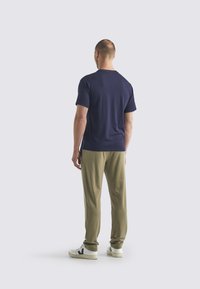 Man som står vänd bort, iförd en marinblå t-shirt, olivgröna byxor och vita sneakers med svarta detaljer mot en ljusgrå bakgrund.