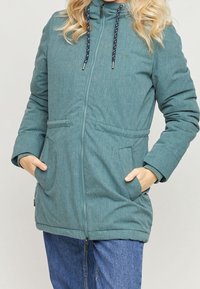 Giacca imbottita teal con colletto alto, chiusura con zip e due tasche frontali. Presenta lacci intrecciati e un tessuto testurizzato.