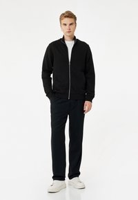 Chaqueta negra con cremallera, camiseta blanca debajo, pantalones negros y zapatillas blancas. Estilo casual con puños y cintura acanalados.