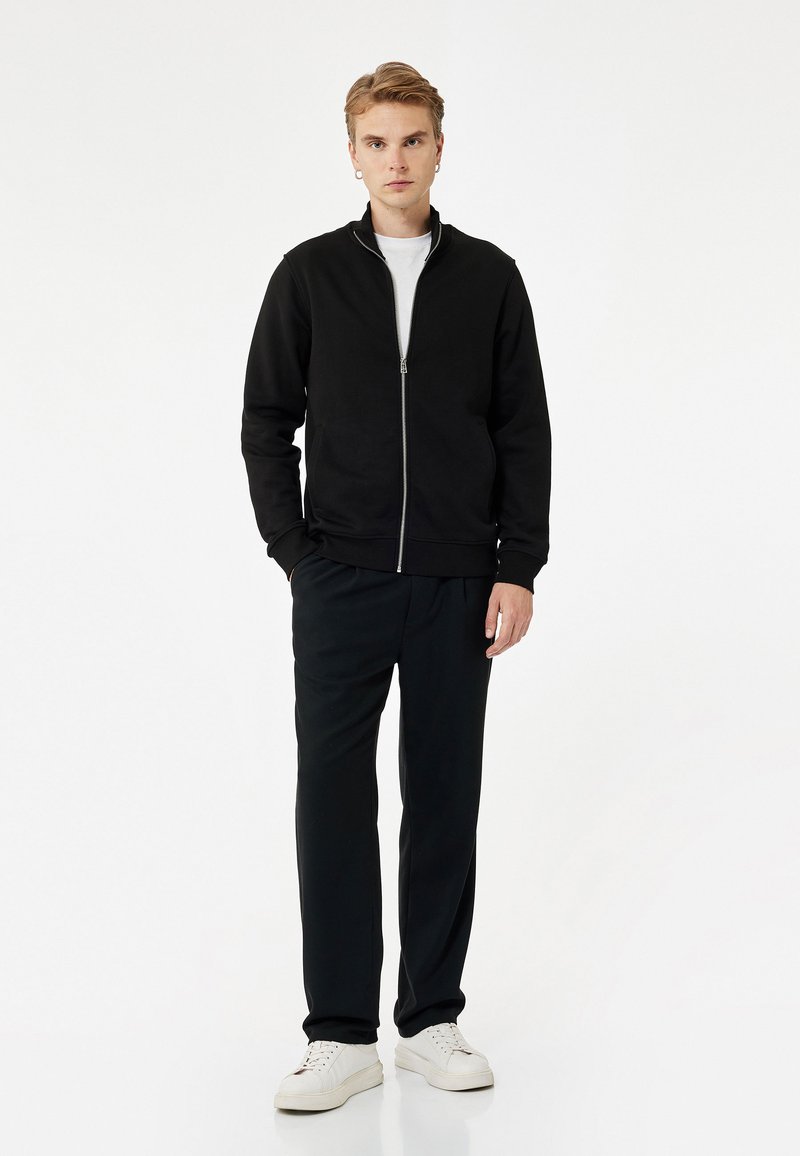 Chaqueta negra con cremallera, camiseta blanca debajo, pantalones negros y zapatillas blancas. Estilo casual con puños y cintura acanalados.