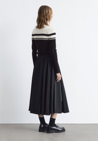 Schwarzer gerippter Pullover mit cremefarbenen und schwarzen gestreiften Akzenten, kombiniert mit einem langen schwarzen Faltenrock. Schwarze Schuhe und Socken runden das Outfit ab.