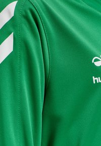 Grøn sportstrøje lavet af glat stof med hvide chevron-striber og logo. Detaljeret syning langs sømmene.