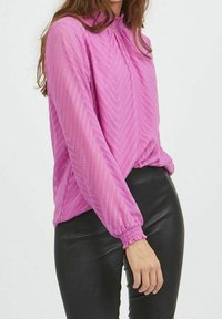 Rosa långärmad blus med hög krage, texturerat zickzackmönster och samlade ärmslut, kombinerad med svarta tajta byxor.