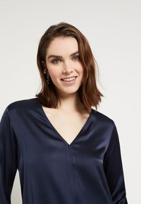 Navy plava satenska bluzica s V-izrezom, dugim rukavima i vertikalnim šavom. Glatka tekstura i suptilna sjajnost poboljšavaju dizajn.