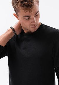 Schwarzes Sweatshirt mit rundem Ausschnitt, langen Ärmeln und einer weichen Textur. Ein geflochtenes Armband ist am Handgelenk sichtbar.