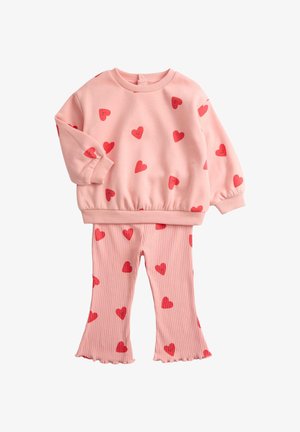 Felpa rosa con stampe di cuori rossi, polsini e vita elasticizzati. Pantaloni rosa coordinati a coste, svasati, con motivi di cuori rossi sparsi.