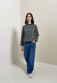 Maglione a maniche lunghe con pattern bianco e nero abbinato a jeans blu larghi e sneakers bianche, in piedi su uno sfondo neutro.