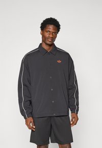 Veste noire avec fermeture à pression, rayures blanches contrastées et logo orange sur la poitrine gauche, avec une coupe décontractée et des poignets élastiques.