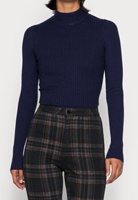 Granatowy sweter z prążkowanym kołnierzem, z długimi rękawami, w zestawie z wysokimi spodniami w kratę w odcieniach czerni, zieleni i brązu.