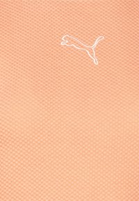 Jasno-brzoskwiniowy materiał z fakturowanym wzorem oraz białym, haftowanym logo Pumy w skaczącej pozycji. Materiał wydaje się oddychający.