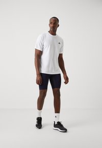 T-shirt de sport blanche avec logo noir, shorts bleus marine et baskets noires. Le modèle porte des chaussettes blanches, au toucher lisse et à la coupe classique.