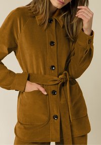 Ruskea corduroy-takki, jossa on kaulus, etunapit, kaksi taskua ja vyötäröllä vyö. Sileä pinta, rento tyyli.