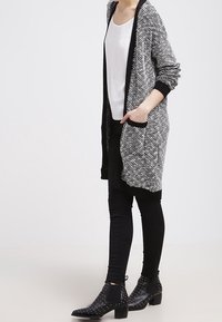 Dlouhý, otevřený černobílý kardigan s texturovaným vzorem, nošený přes bílé tričko, spárovaný s černými skinny džíny a kotníkovými botami s cvočky.