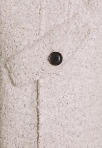 Vitt texturerat tyg med ett vävt mönster. Innehåller en svart plastknapp med fyra hål och en sidoficka.