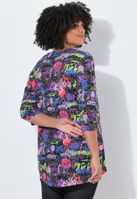 Frau mit lockigem Haar, die ein langärmliges Shirt mit buntem Graffiti-Muster trägt, seitlich steht und eine Hand hinter ihrem Rücken hat.