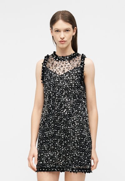 Jeune femme portant une mini robe noire sans manches avec un haut transparent et des sequins argentés partout, debout devant un fond uni.