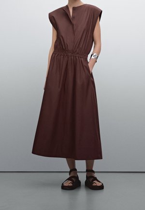 Brown, sleeveless dress with a buttoned neckline and elastic waist. The fabric appears smooth, and the silhouette is long and loose-fitting.