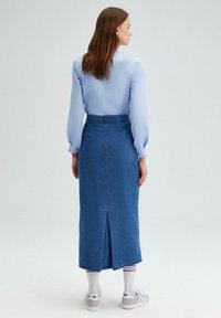 Jupe en denim délavé, longueur cheville avec une fente à l'arrière, associée à un chemisier bleu clair et des baskets blanches, vue de dos.