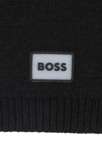 Beanie nero a maglia con bordo a coste e una toppa rettangolare bianca con "BOSS" in caratteri neri bold. Texture morbida.