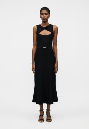 Kobieta stojąca w czarnej, bezrękawowej sukience midi z wycięciem o skręconym dekolcie i cienkim czarnym paskiem, ubrana w czarne sandały na paski.